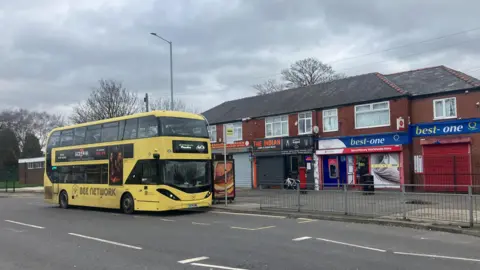 Photograph of a Bee Network bus on the Brinnington estate in Stockport. The image also shows a parade of shops.