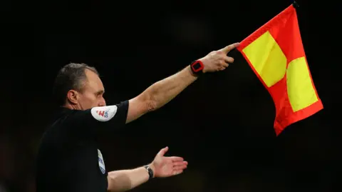 Getty Images Darren Cann, an assistant referee who is officiating a match. He can be seen in black uniform and is waving a yellow and red flag.