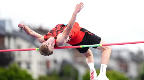 PA Media Image shows high jump contestant Regan Corrin, in black shorts and red top, jumping over a red bar