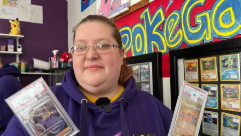 BBC Toni Marlow has a purple jumper and is wearing glasses, she is holding two Pokémon cards and is stood in front of a wall with Pokémon merchandise in the background.