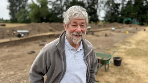 Josh Gorrono-Chapman/BBC Dr Peter Halkon pictured standing next to an area that has ben excavated. He has grey hair and a grey beard and is wearing a brown fleece and grey polo shirt.