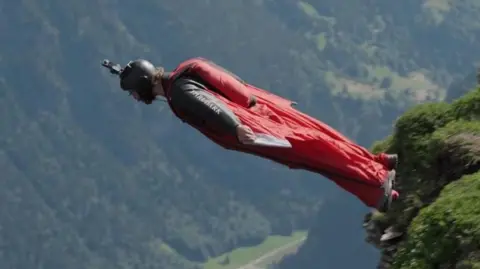 Liam Byrne in red wing suit flying through the sky. Picture taken from 2024 BBC Scotland documentary