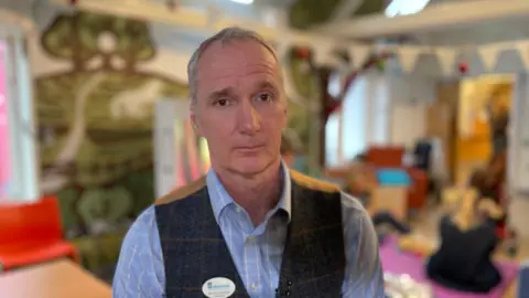 A man standing in a colourful play room wearing a pale blue shirt and checked tweed brown and cream waistcoat with a solemn expression on his face