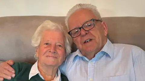 Luke Deal/BBC Jo Johnson and Terry Smith sit together on a sofa. Jo has short grey hair and wears a green jumper with a white shirt underneath. Terry has short grey hair, glasses and a blue shirt. He has his arm around Jo.