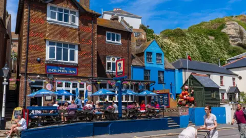 The pub "The Dolphin Inn" on Rock-a-Nore Road in Hastings on a sunny day. Some people are dining at the outdoor tables, others walking on the pavement. The cliffs rise in the background.