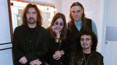 Butler, Osbourne, Ward and Iommi, dressed in black, sitting around a lit dressing room mirror. Their expressions are solemn as they look at the camera.