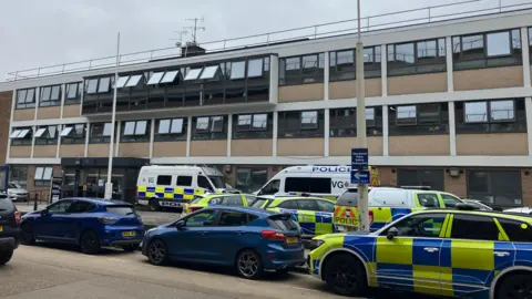 Henry Godfrey-Evans/BBC Several police vehicles parked outside a large building