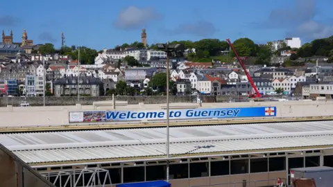 Photo of the roof of Guernsey harbour with a 'Welcome to Guernsey' sign. There are houses and buildings in the background and a blue sky.