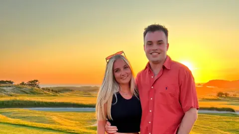 Family photo Jack and Emily holding each other and smiling for the camera. Jack is wearing a pink button down t shirt and has one arm around Emily's waist. He has short cropped brown hair. Emily is wearing a fitted black sleeveless top. She has straight blonde hair down below her shoulder and has sunglasses on the top of her head. They are standing on groomed grass in front of a golf bunker with sun setting in the background