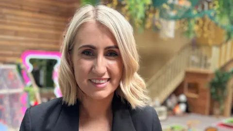 Laura McManus tiene el pelo rubio corto y está sonriendo. Lleva una chaqueta negra.
