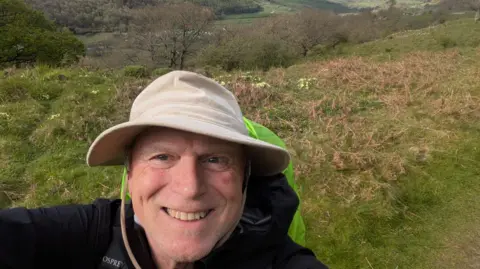 Alan Chape Alan Chape is wearing a cream sun-hat and has on a backpack while he walks along a grassy path. A valley can be seen in the distance behind him.
