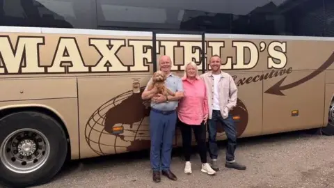 Man in jeans, a shirt and holding a dog, standing next to a blonde woman with a pink shirt, black leggings and trainers and a young man in a shirt and jeans, standing in front od a brown bus with 'Maxfield's Executive'