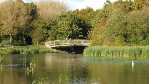 Paul Bryan/ Geograph Wooden footbridge over a waterbody- surrounded with green trees and grass- with a white swan in the water