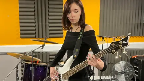 A young woman playing an electric bass guitar with a drum kit behind her.