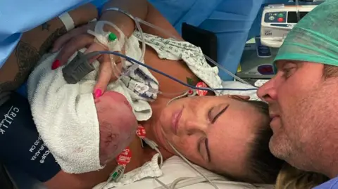 A woman holding a newborn baby while attached to a number of wires. A man wearing a green mesh cap is crouched next to her.