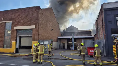 Seven firefighters dressed in brown firefighting uniforms with yellow helmets work at the scene of a fire. Smoke is billowing into the sky behind a building in the background. Yellow hoses from a fire truck on the right can be seen snaking along the ground and a ladder has been placed against a roof in the background.