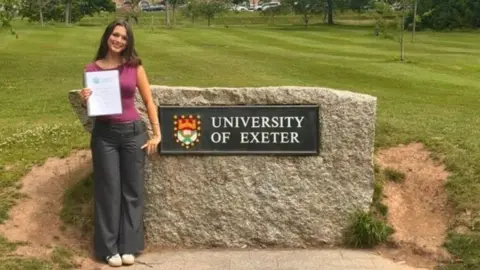 Mia Taylor-Seal A young woman with brown hair is wearing a pink top and black trousers. She is holding her dissertation in her hand and is standing next to a large sign that reads "university of essex"