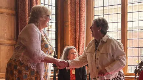 Anne Hope and Jenny Watkins shaking hands in an ornate, wood-panelled room. A woman in mayoral chains is sat in a chair behind them. The women are smiling.