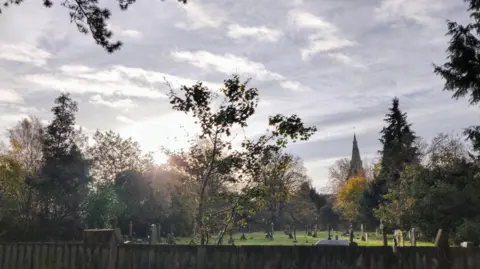 Georgeta An early sunrise can be seen with the sun coming up behind a row of trees. A short fence can be seen at the front of the image with a graveyard behind. A church spire can also be seen behind some of the trees.