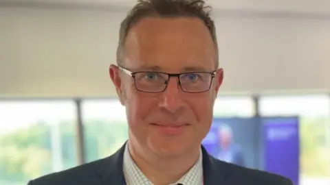 Supplied Richard Taylor, wearing a shirt, blazer jacket and glasses, looks straight ahead.