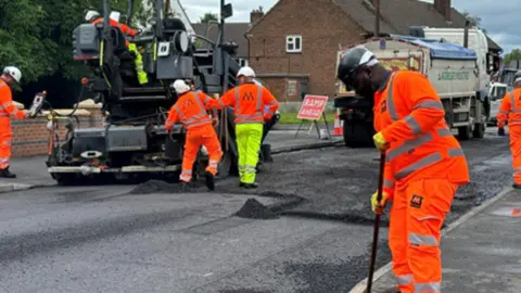 Staffordshire County Council Road workers wearing orange and yellow hi-vis jackets and trousers work in a road, with large vehicles to their left and a building behind them. There is a sign behind them reading "ramp ahead"