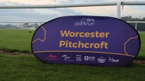 Worcester Pitchcroft Parkrun A purple oval sign that is upright on the grass and reads "Worcester Pitchcroft" in orange writing. There is a white railing above the sign.