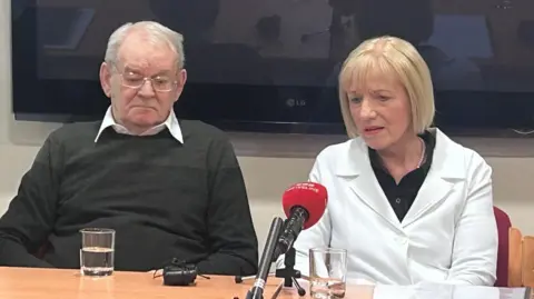 Alan Black has short grey hair and is wearing metal framed glasses, a green jumper and a white shirt. He is looking down at the table. Karen Armstrong has short blonde hair and is wearing a white blazer with a black shirt and has pink lipstick on. She is looking at the red BBC news mic. They are sitting down a wooden table which has glasses of water, a mic and paper on it.