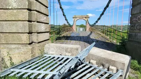 The photo has been taken from one side of an old style suspension bridge. The floor is wooden, and curves in the distance as it reaches the other side. There are two barriers at the front of the bridge that have been knocked down. The sky is blue.