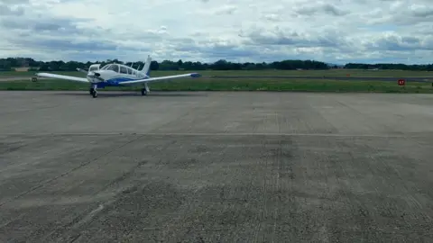 CARMELO GARCIA A small private propeller plane sits on the tarmac at Gloucestershire Airport. It is blue and white and more runway is visible in the background. 