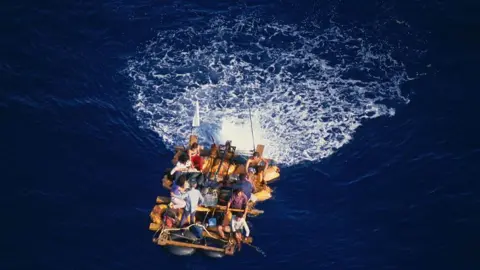 Getty Images A group of people on a makeshift raft sail on the way, with a white wake behind them