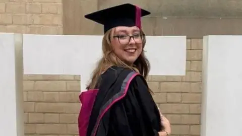 Stephanie Irons wearing a graduation gown and cap in front of a sign for Nottingham Trent University