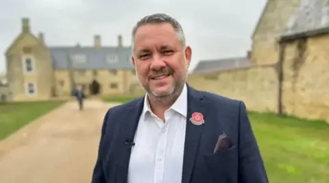 Kate Bradbrook/BBC Jason Smithers with greying hair and a beard wearing a blue suit and a white open necked shirt. He is standing in front of a yellow brick building of older construction.