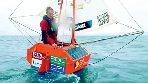 Andrew Bedwell, in his early 50s, stands in a tiny orange yacht on the open ocean, which has a white sail and is covered with signs for various sponsors. He is wearing a black life vest and red waterproofs.