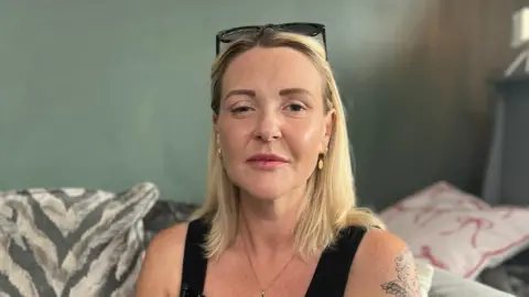 Nicola Fairly, a blonde woman in her 30s who is wearing a black vest and black sunglasses on her head. One side of her face is droopy which is a symptom of botulism. She has a tattoo on her left shoulder and is sitting on a sofa with cushions of different patterns.