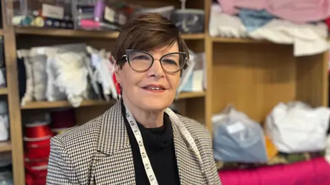Shirlee Meads, stood in front of shelves of lace, material and ribbon, looking at the camera