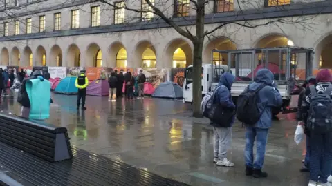 MancStPauli A small van is parked in front of the arches at St Peter's Square as council workers throw red tents into a trailer on the back. Three men wearing hoodies and rucksacks are seen watching on. A group of other people surrounded one of the tents 