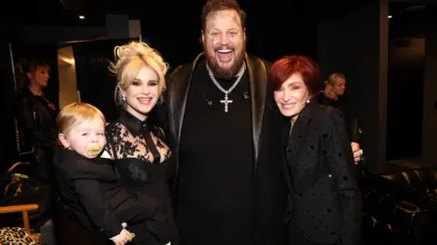 Getty Images Kelly, filha de Ozzy, tem cabelos loiros e usa um vestido de renda preto. Ela está segurando seu filho pequeno, Sidney. Um homem, Jelly Roll, veste uma jaqueta de couro e um crucifixo. Ele está sorrindo, tem tatuagens no rosto, cabelo castanho curto e barba. Ele está com o braço em volta de uma mulher, Sharon Osbourne, que tem cabelos ruivos e veste um casaco preto. Ela está sorrindo.