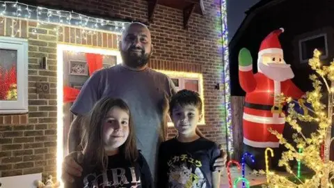 George Carden/BBC Jose De Sa wears a grey t-shirt and stands in front of his decorated house with his two children, a boy and a girl, both wearing dark t-shirts.