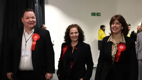 EPA Labour candidate Angeliki Stogia and Labour Party Deputy Leader Lucy Powell smile and walk alongside a male party worker as the enter the hall where results were declared. They all wear dark suits with red party rosettes.