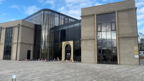 A large mosque with a major central structure made of glass and two brick built towers either side. The front is paved with a number of cones in place 