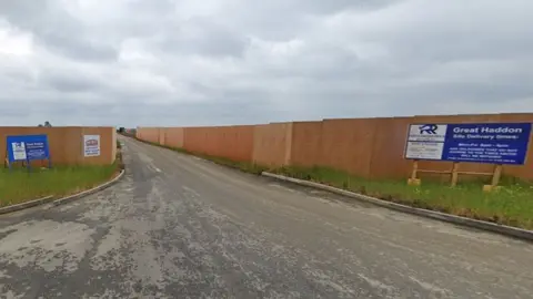 Google Entrance road to a building site, with tall beech coloured wooden fence panels on either side. A blue sign on the right side says Great Haddon Site Delivery times, and there is a blue notice on the left side with white writing. The grey road is rough and a bit bumpy.