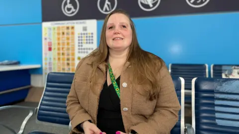 Emilie Douglas smiles as she sits on a blue arm chair and in the background you can see the special assistance area at Jersey Airport