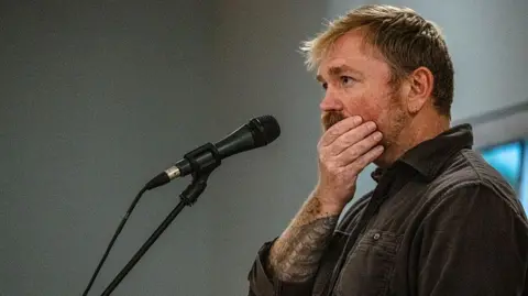 Graham Platner seen on stage at a microphone. his hand is over his mouth 