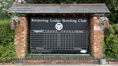 Ollie Conopo/BBC A black scoreboard at a bowling club, with a roof above, made of grey slate, and red bricks all around it. Hanging baskets are either side and bushes on both sides. 
