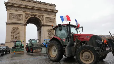 Several tractors, including a red tractor flying French flags, are parked in front of the Arc de Triomphe in Paris.