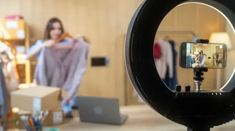 Getty Images A content creator holds up a cardigan on a hanger while facing at a smartphone recording her on a stand with a ring light.