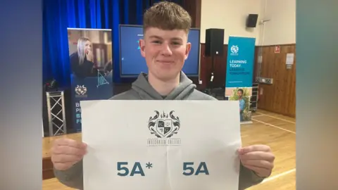Boy with brown hair and grey hoodie. Holding sign saying 5A* and 5As