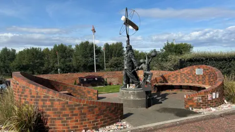 Outdoor memorial featuring two bronze figures on a circular stone pedestal, surrounded by curved red-brick walls and semi-circular benches. One figure raises a large metal structure overhead while the other stands nearby. The memorial sits in a grassy area with trees and flagpoles. It a sunny day with blue skies and some clouds.
