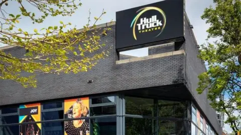 Exterior of Hull Truck Theatre with black brick wall and large glass windows with the theatre's name on a sign at the top corner. Posters for upcoming plays hang in the window.