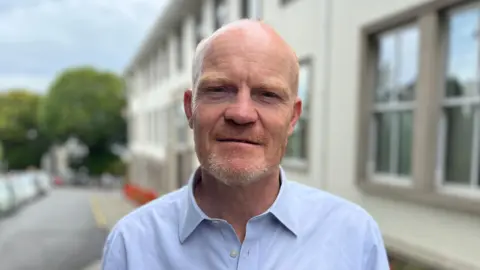 Deputy Gavin St Pier is pictured. He is a man with a bald head looking at the camera. He has a ginger and grey beard. He is wearing a blue shirt. 